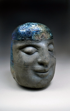 Statue of a head, about the size of a human head, in the ancient style of the Olmec civilization. Mostly dark colored from smoky firing, but with blue iridescence in a band across the forehead.
